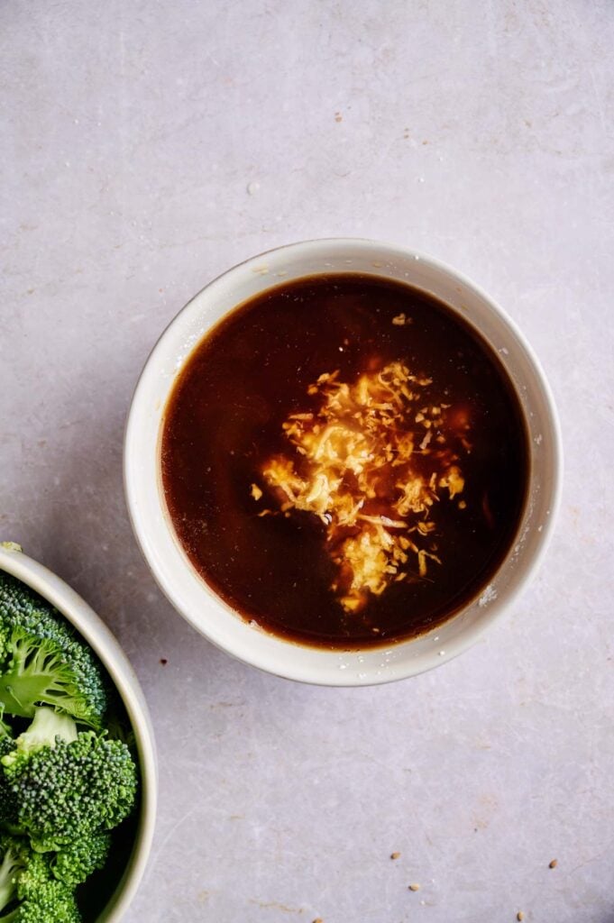 A bowl of brown sauce with grated cheese or garlic on top, next to a partial view of a bowl with broccoli.