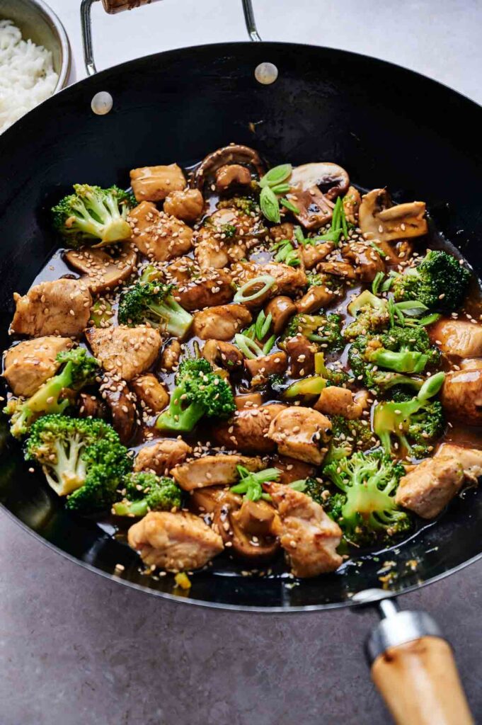 Stir-fry dish in a wok featuring chicken, broccoli, mushrooms, and green onions, garnished with sesame seeds.