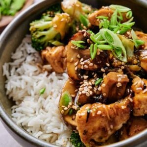 A bowl of rice topped with stir-fried chicken, broccoli, and mushrooms, garnished with sesame seeds and chopped green onions.