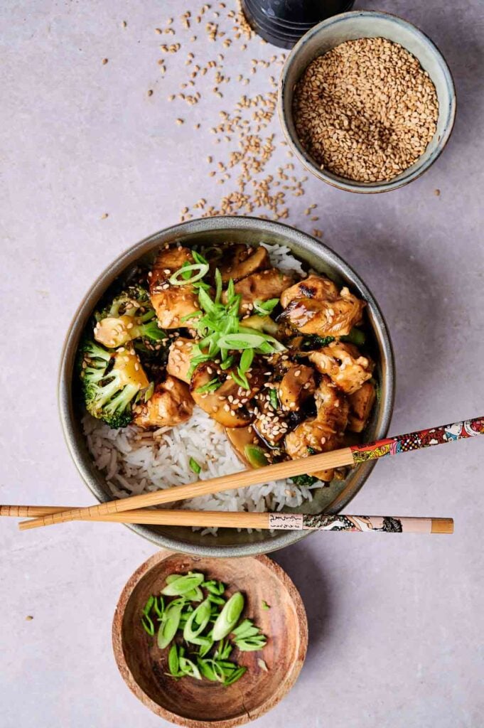 Bowl of rice topped with chicken, broccoli, and green onions, paired with chopsticks. Small bowls of sesame seeds and sliced green onions are nearby.