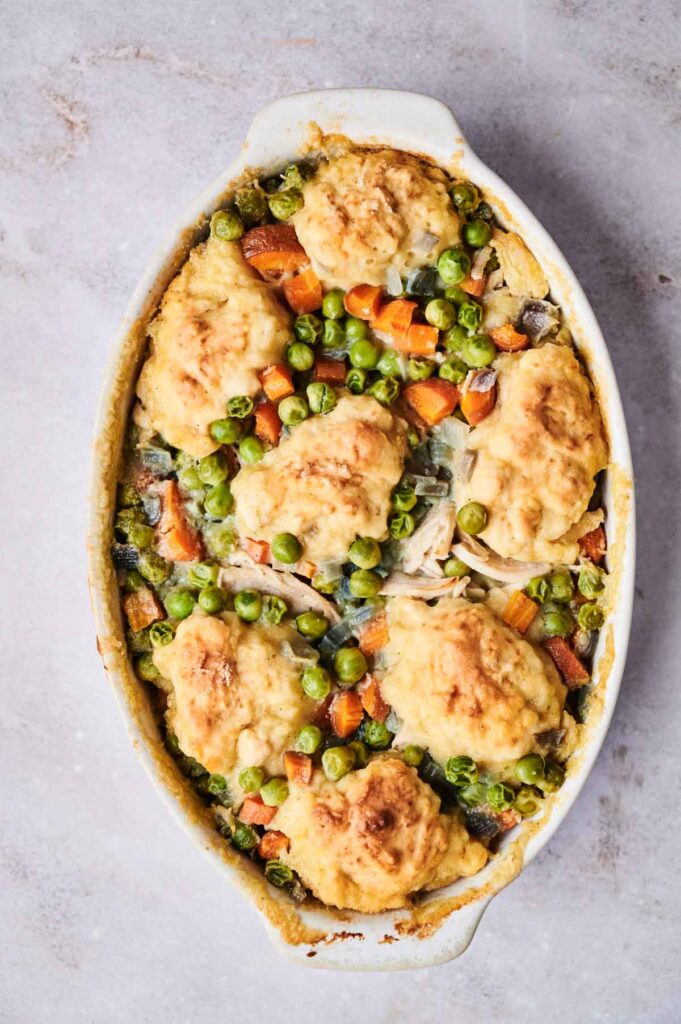 An oval dish filled with a baked chicken pot pie, topped with golden brown biscuits, visible vegetables including peas, carrots, and onions.