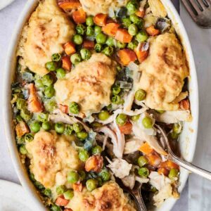 A baked dish with chicken, peas, carrots, and dumplings in a cream sauce, served in a white oval casserole dish with two silver forks.