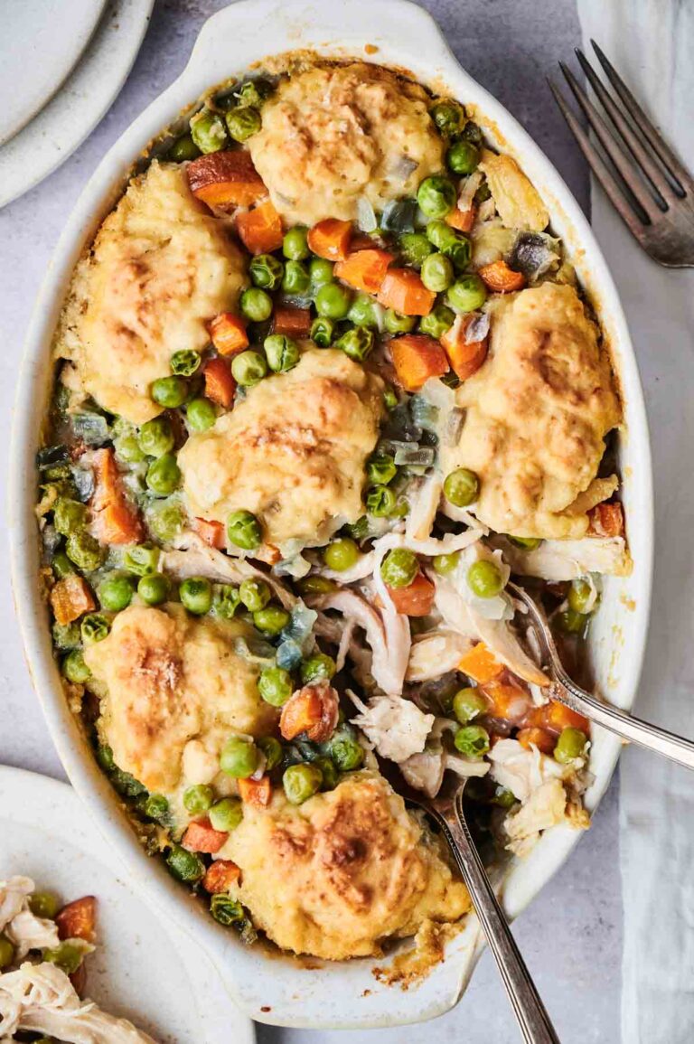 A baked dish with chicken, peas, carrots, and dumplings in a cream sauce, served in a white oval casserole dish with two silver forks.