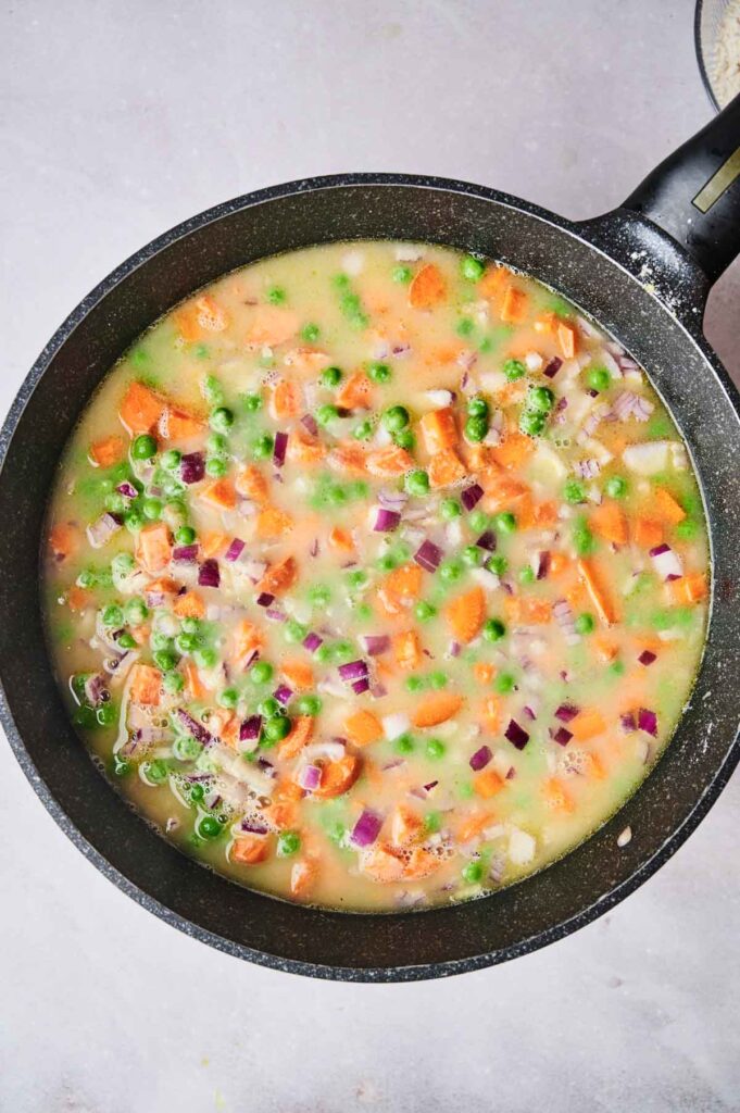 A pan filled with a creamy mixture of chopped carrots, peas, red onions, and other vegetables, ready for cooking.