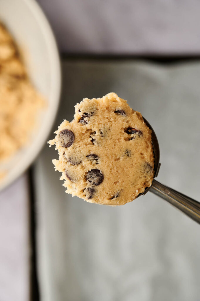 Close-up of a spoonful of chocolate chip cookie dough, with visible chocolate chips, held over a baking tray.