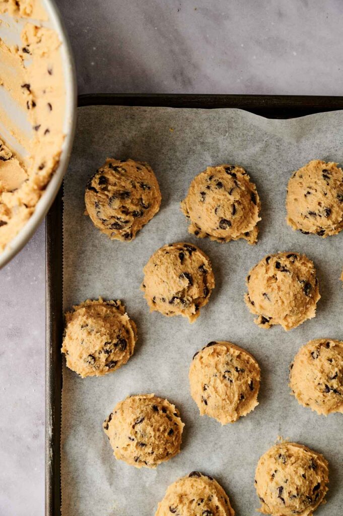 Balls of chocolate chip cookie dough on a parchment-lined baking sheet next to a mixing bowl.