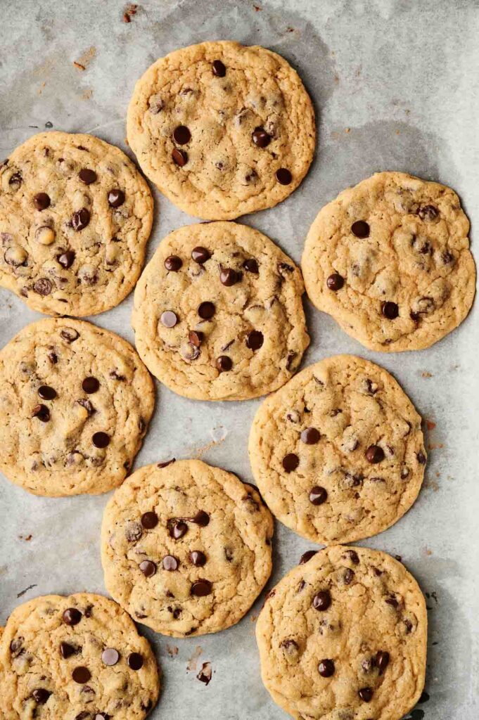 Ten chocolate chip cookies on parchment paper, arranged in a scattered pattern.