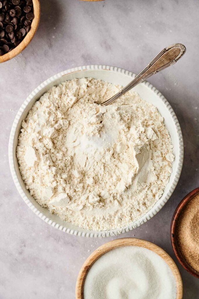A bowl of flour with a spoon, surrounded by small bowls of chocolate chips, sugar, and brown sugar on a marble surface.