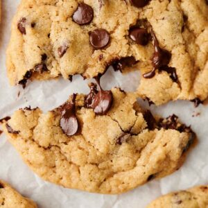 A close-up of warm, gooey chocolate chip cookies on parchment paper, with one cookie broken in half, revealing melted chocolate.