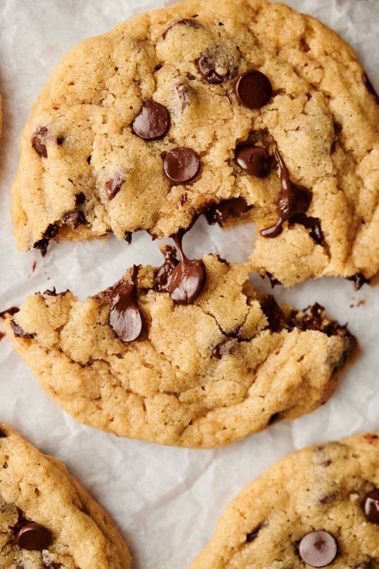 A close-up of warm, gooey chocolate chip cookies on parchment paper, with one cookie broken in half, revealing melted chocolate.