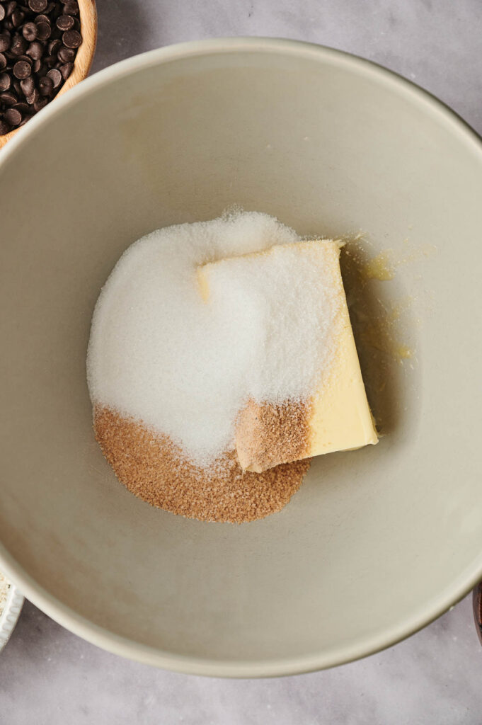 A mixing bowl with butter, white sugar, and brown sugar on a gray surface.
