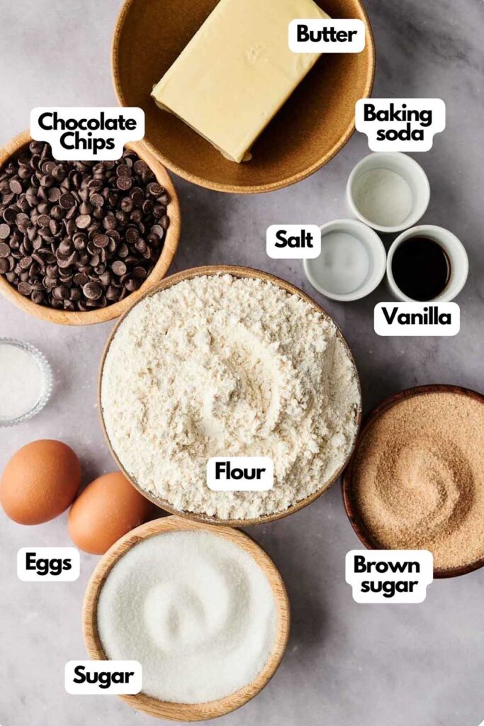 Baking ingredients arranged on a table include flour, eggs, butter, sugar, brown sugar, chocolate chips, baking soda, salt, and vanilla.