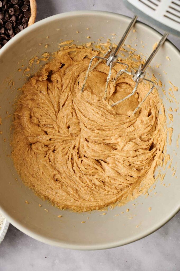 A mixing bowl containing brown sugar cookie dough, with a hand mixer partially inserted. Chocolate chips are visible beside the bowl.