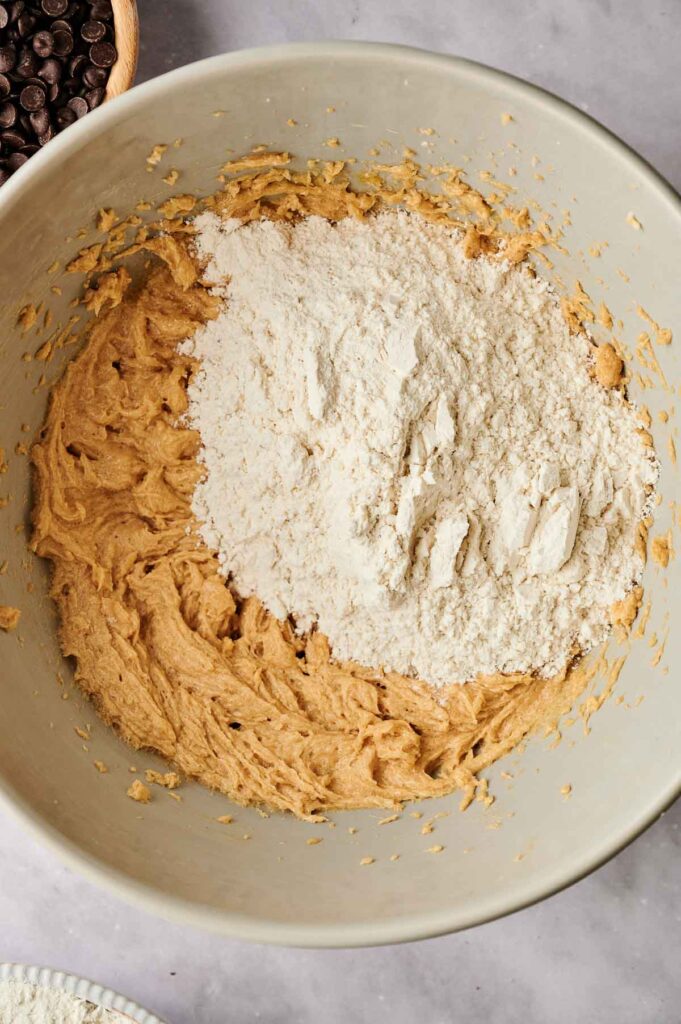 Bowl with cookie dough mixture, partially combined with flour on top. Nearby are chocolate chips and a glimpse of a white ingredient.