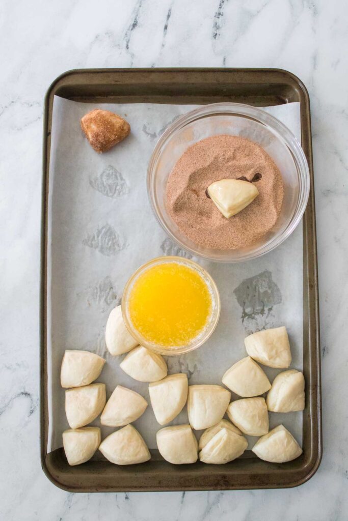 A baking tray with cinnamon sugar biscuit bites, dough pieces glistening with melted butter, and a bowl of sweet spice sits on a parchment paper-lined surface.