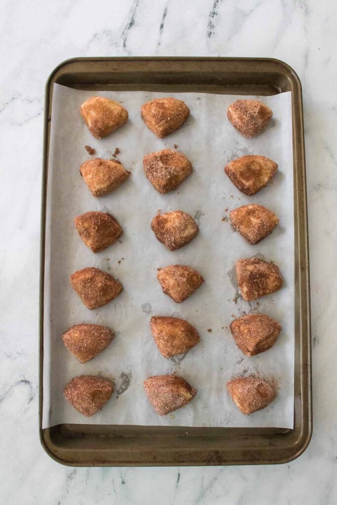 A baking tray with 18 evenly spaced Cinnamon Sugar Biscuit Bites on parchment paper, each coated in a sweet layer of cinnamon sugar, set on a marble surface.