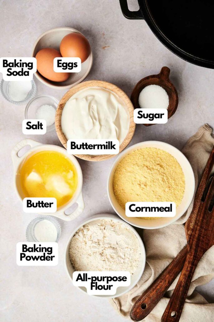 Labeled ingredients for Air Fryer Cornbread: eggs, baking soda, salt, buttermilk, sugar, melted butter, cornmeal, baking powder, and all-purpose flour are neatly arranged on a countertop.