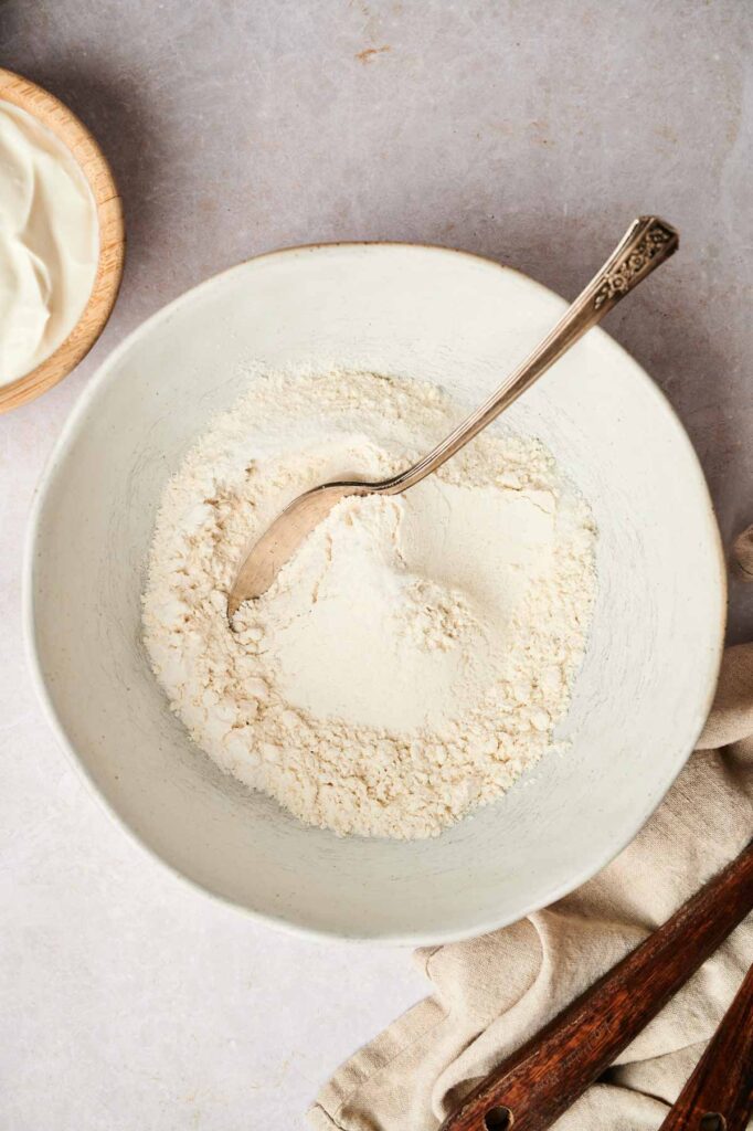 A bowl of flour with a metal spoon on a gray surface, ready to craft delicious air fryer cornbread. A wooden bowl and a beige cloth are partially visible, setting the scene for culinary creativity.