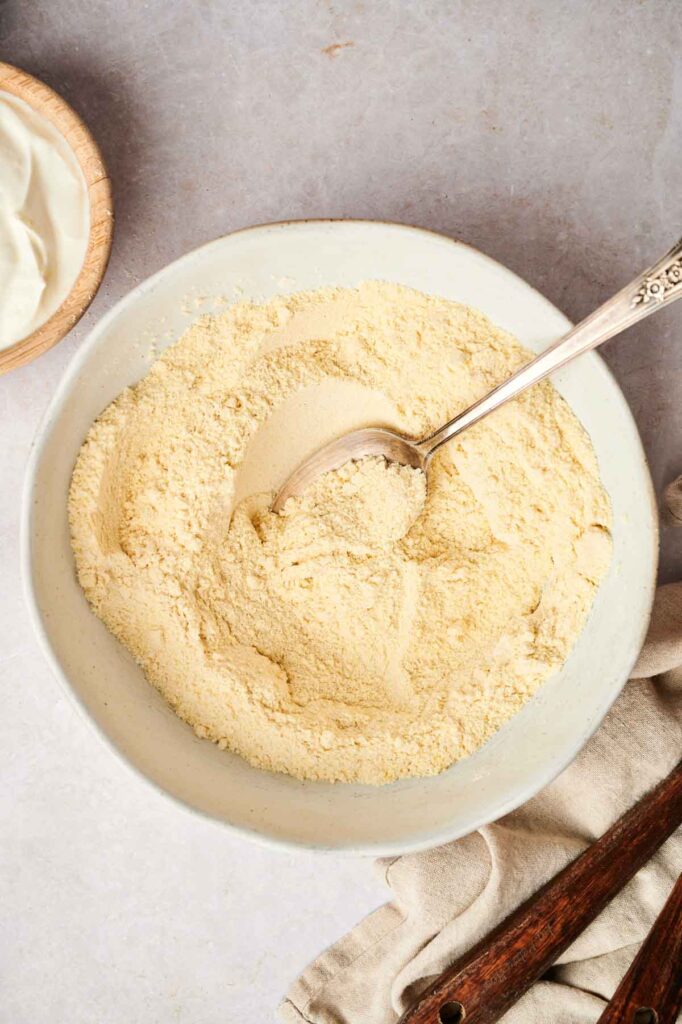 A bowl of yellow cornmeal with a metal spoon on a gray surface sits ready, suggesting the beginnings of air fryer cornbread, accompanied by a wooden bowl and cloth.