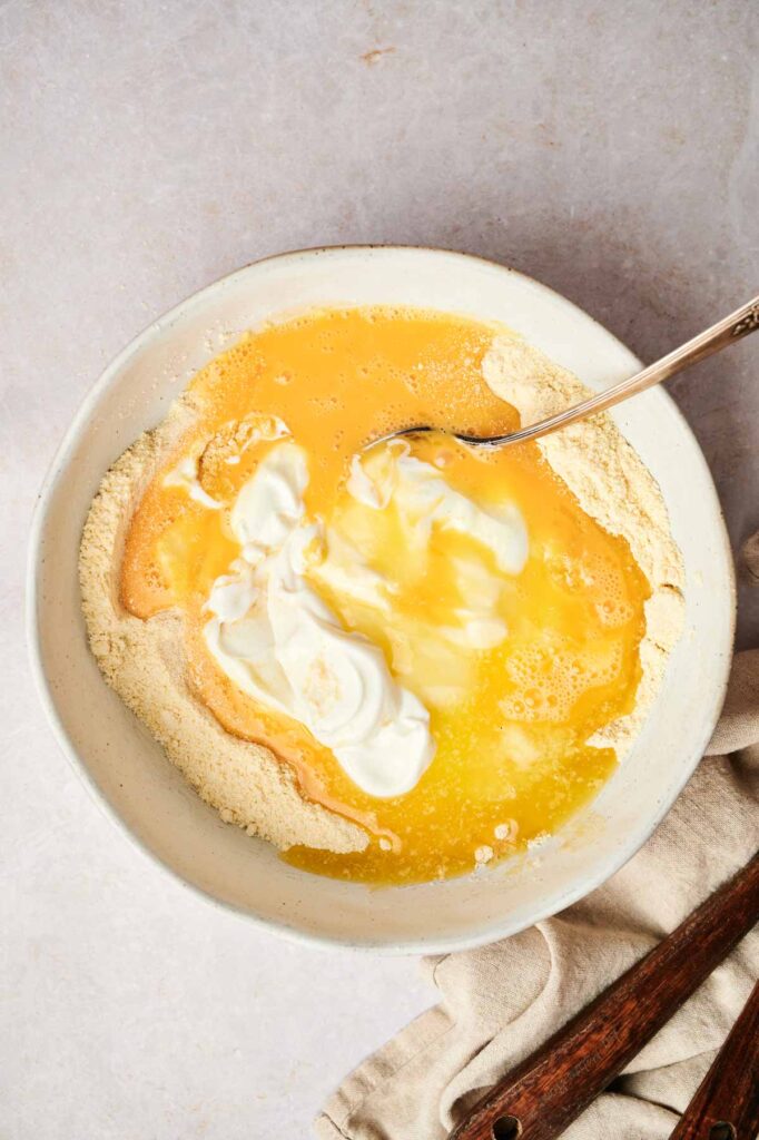 A bowl with flour, beaten eggs, and dollops of cream is mixed with a spoon to create a delightful air fryer cornbread batter.