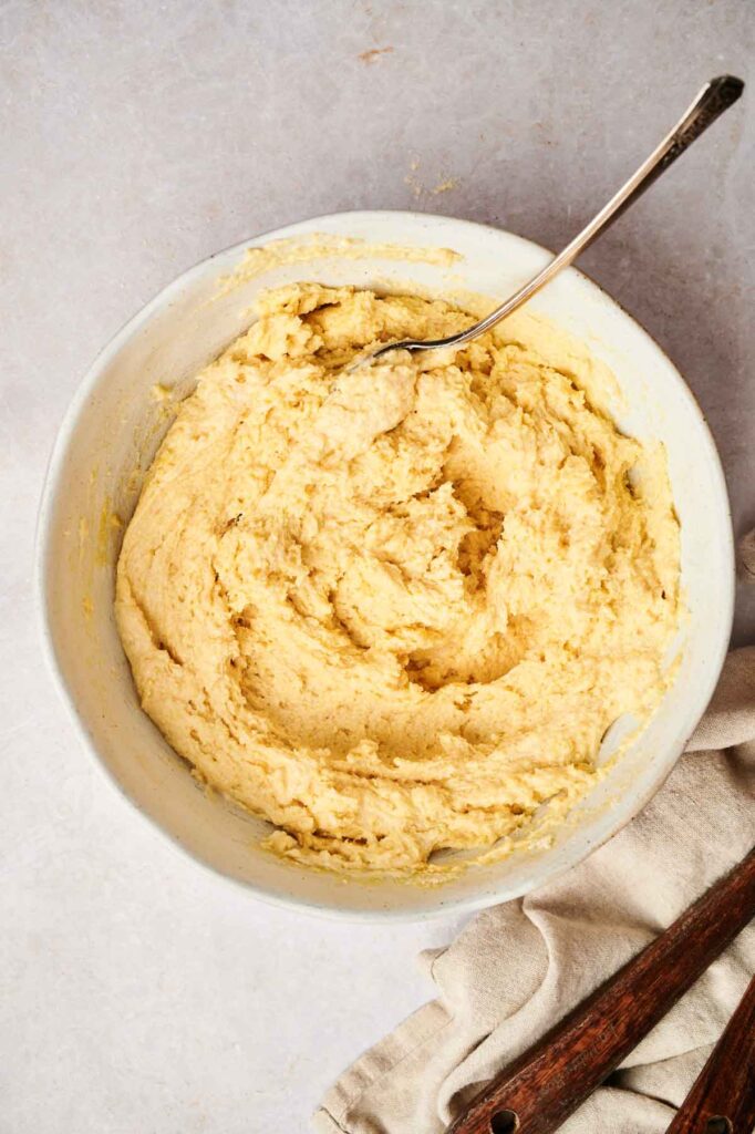 A bowl of creamy yellow cookie dough rests on a light surface, paired with a metal spoon and a beige cloth nearby. Imagine the aroma mingling with the warm scent of air fryer cornbread wafting through the kitchen.
