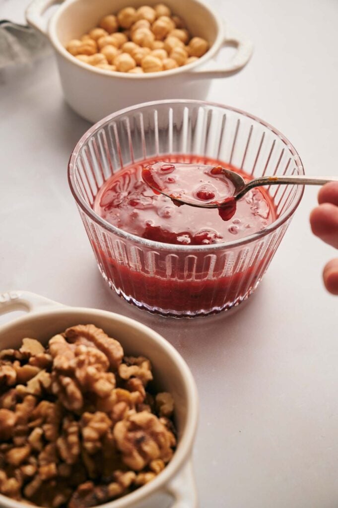 A glass bowl of red jam with a spoon, surrounded by small dishes of hazelnuts and walnuts.