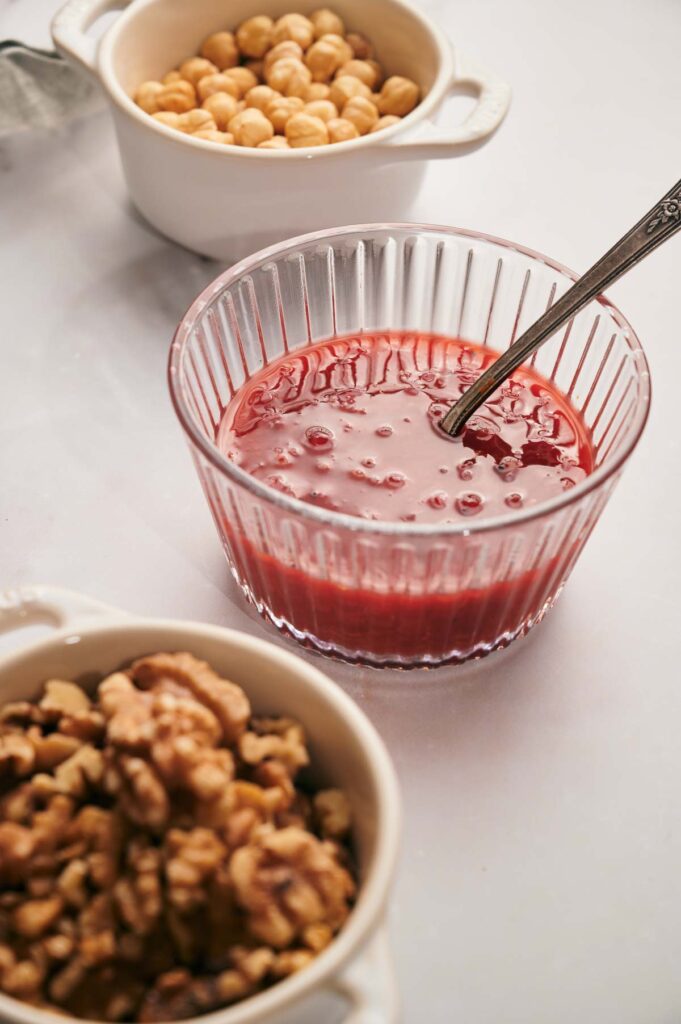 Glass bowl of red sauce with a spoon, surrounded by a white bowl of chickpeas and another of walnuts on a light surface.