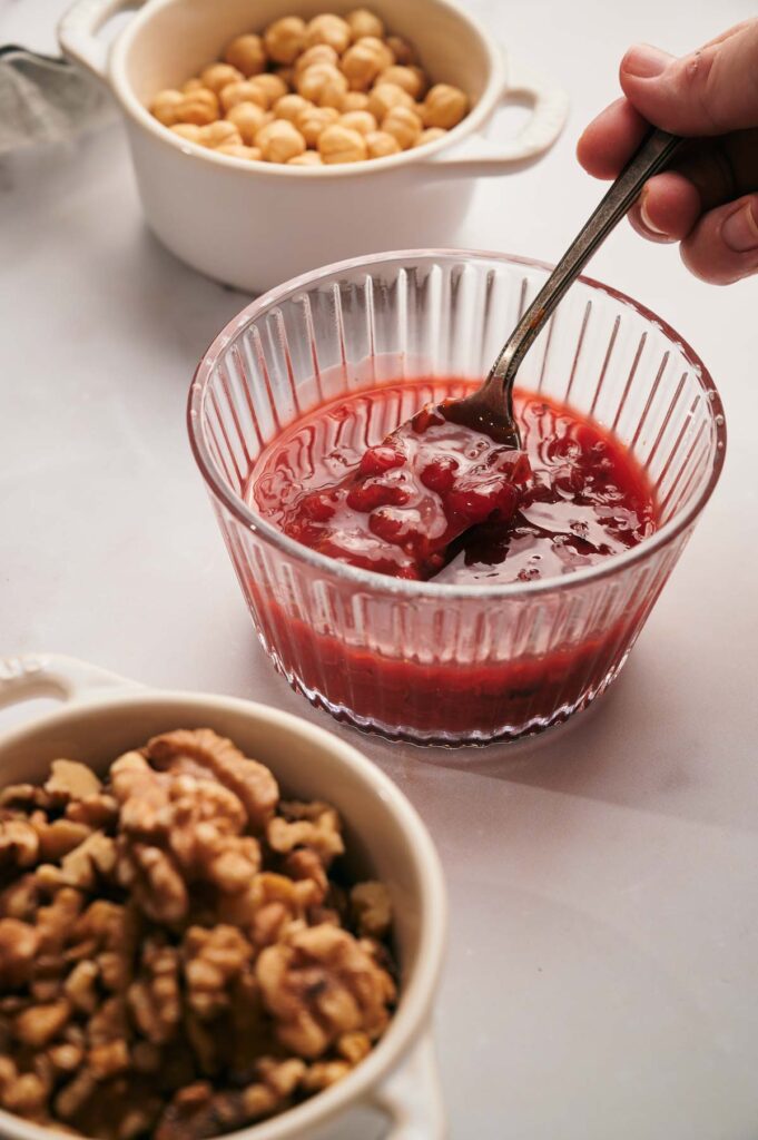 A hand holds a spoon in a bowl of red jam, with separate bowls of chickpeas and walnuts nearby on a light surface.