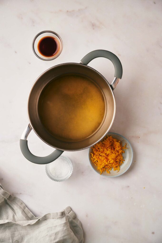 A pot with liquid on a light surface, surrounded by small bowls containing a dark liquid, grated orange zest, and salt. A cloth is partially visible at the bottom left.