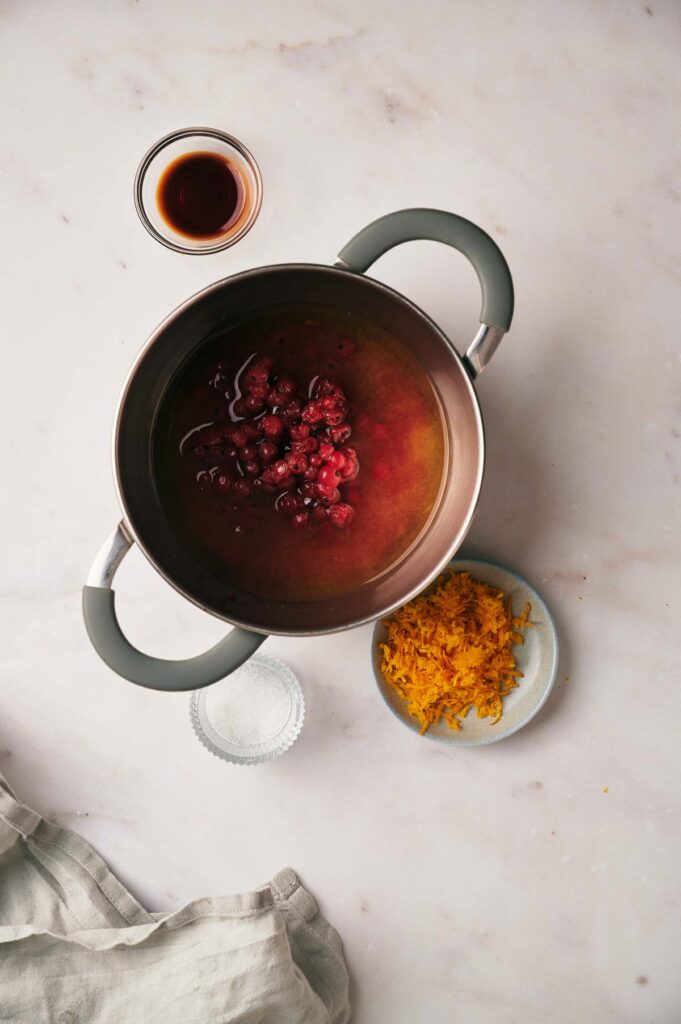 A pot with a red liquid and berries inside, a small bowl of vanilla extract, grated orange zest on a plate, and some sugar on a light marble surface.