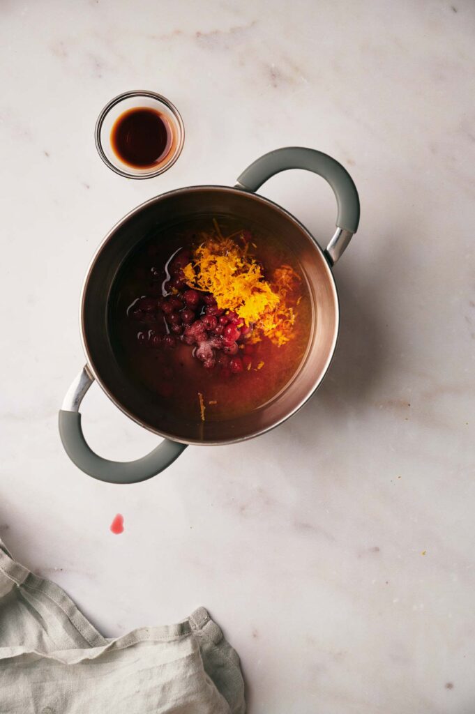 A pot with cranberries, orange zest, and liquid. A small bowl with a brown liquid is nearby on a marble surface.
