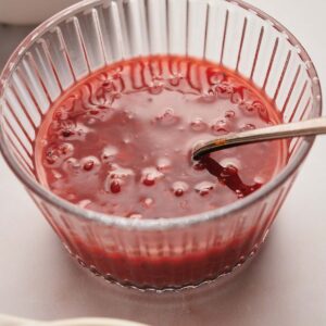 A clear glass bowl filled with red jelly containing small berries, with a spoon resting inside.