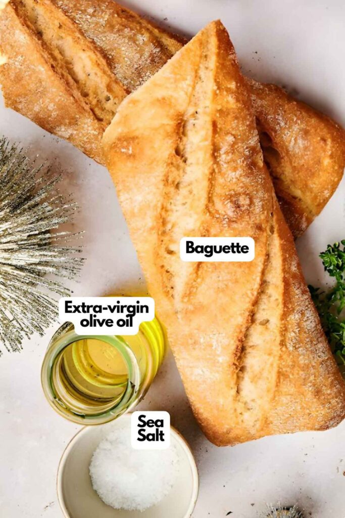 Two baguettes on a surface with a bowl of extra-virgin olive oil and a bowl of sea salt nearby, alongside a sprig of greenery.