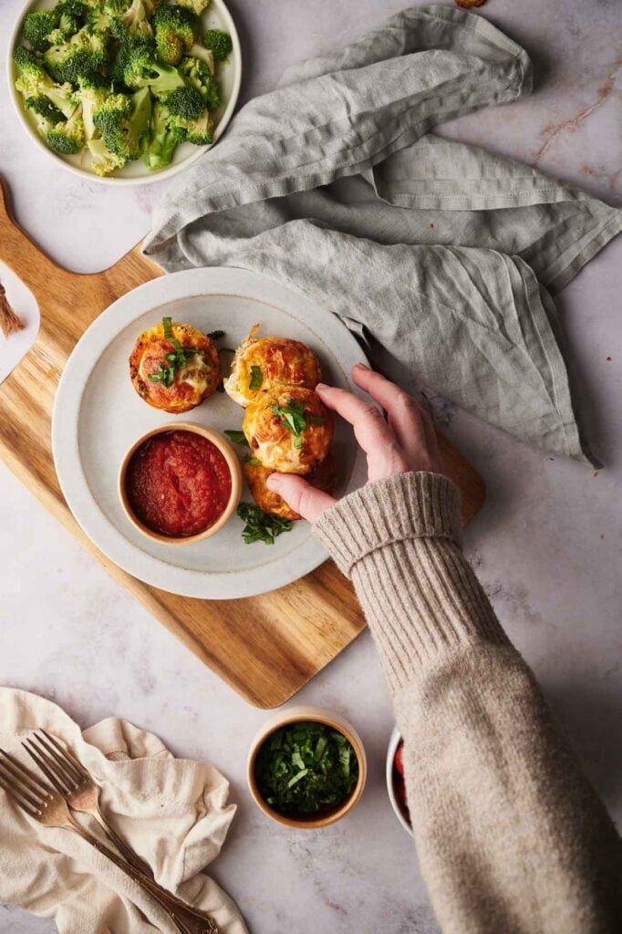 Hand reaching for a plate with three stuffed bell pepper halves and a bowl of tomato sauce on a wooden board, accompanied by bowls of broccoli and chopped herbs. A gray cloth napkin is nearby.