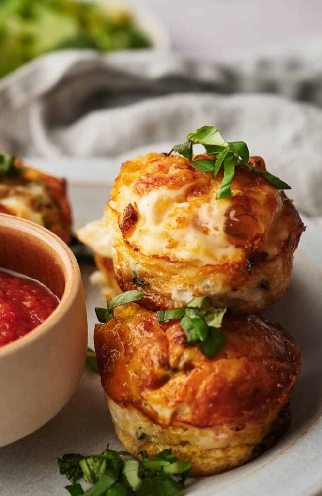 Two golden-brown egg muffins topped with fresh herbs on a plate, next to a small bowl of red sauce.