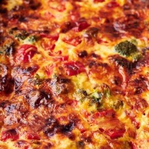 A baked casserole with a golden-brown top, featuring visible vegetables such as broccoli and tomatoes, in a rectangular dish with handles.