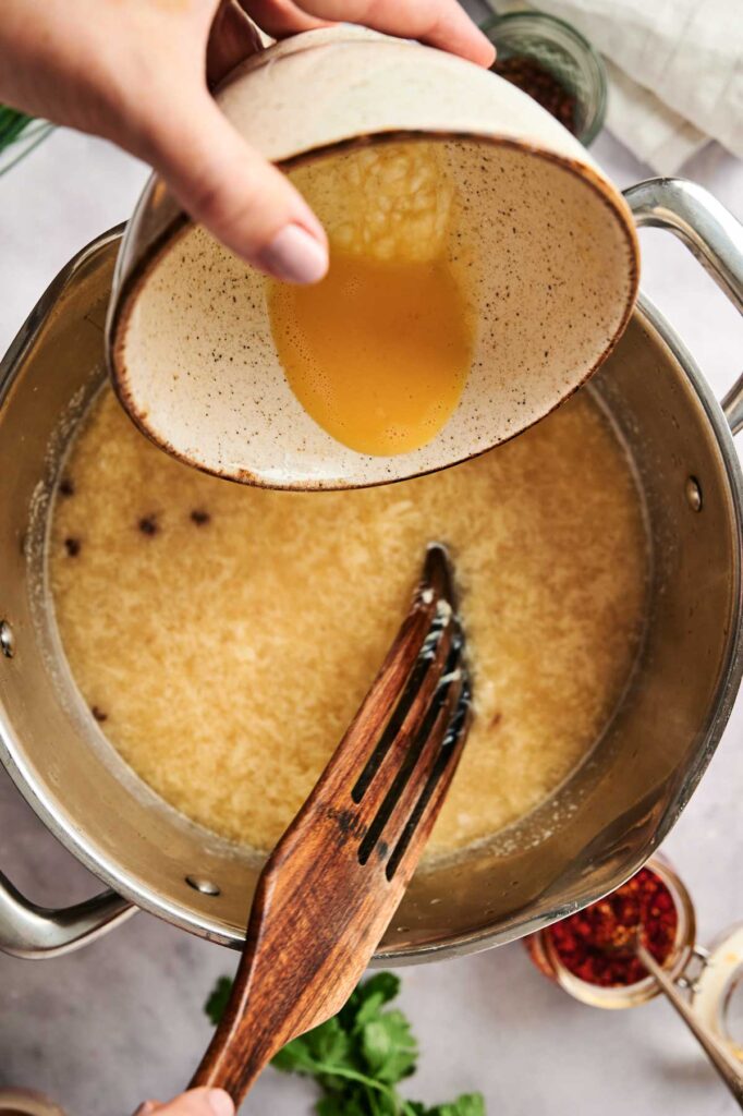 Hands pouring liquid from a bowl into a pot, with a wooden spatula stirring. A delightful aroma fills the air as egg drop soup ingredients are visible in the background, promising a comforting meal.