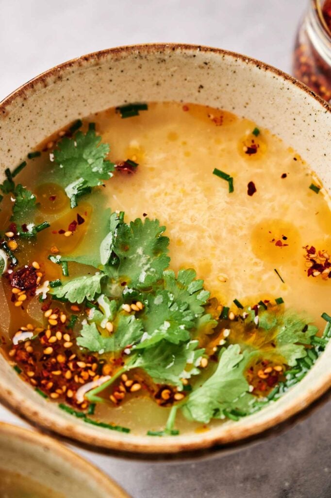 A steaming bowl of delicious egg drop soup, delicately garnished with fresh cilantro, chives, and a sprinkle of red chili flakes.