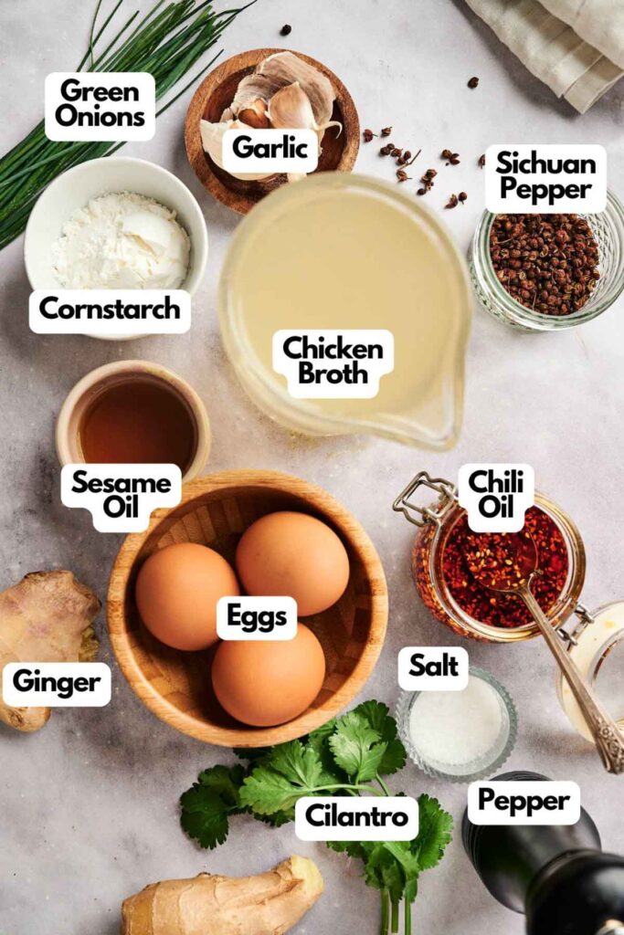 A variety of labeled ingredients for cooking egg drop soup, including green onions, garlic, Sichuan pepper, cornstarch, chicken broth, sesame oil, eggs, chili oil, ginger, salt, cilantro, and pepper.