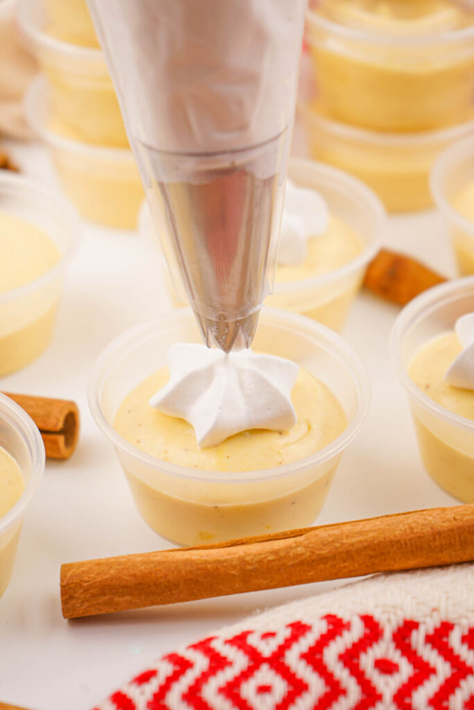 Piping white cream onto dessert cups filled with eggnog pudding shots, surrounded by cinnamon sticks and a red patterned cloth.