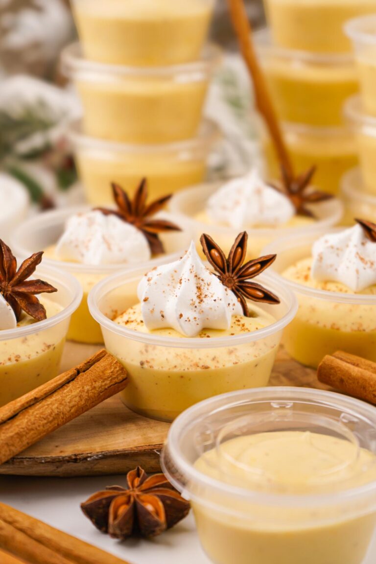 Several small cups of flan topped with whipped cream and star anise are artfully surrounded by cinnamon sticks, evoking a festive feel akin to eggnog pudding shots. In the background, more flan cups are stacked, ready to delight any dessert lover.