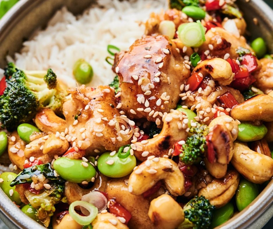 A bowl of rice topped with cashew chicken, featuring stir-fried broccoli, red peppers, edamame, green onions, and sesame seeds.