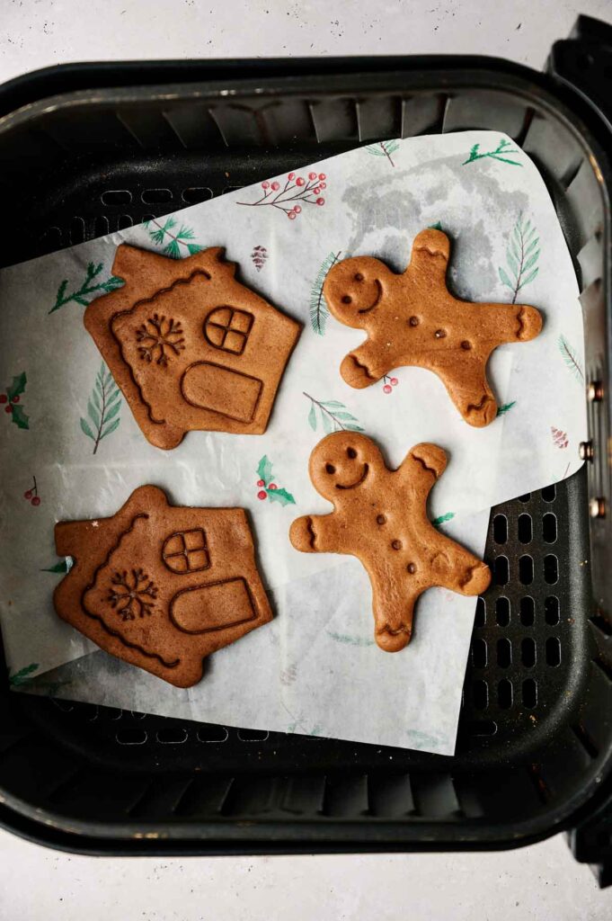 Gingerbread cookies shaped like houses and people rest perfectly on parchment paper, nestled in an air fryer basket. These delightful air fryer gingerbread cookies offer a festive twist with crisp edges and warm spice aromas that fill the room.