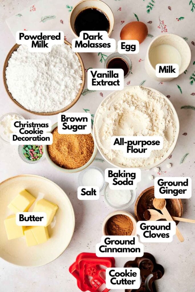 Baking ingredients for air fryer gingerbread cookies laid out: powdered milk, molasses, egg, milk, vanilla, flour, brown sugar, baking soda, spices, butter, and cookie cutter with holiday decorations.