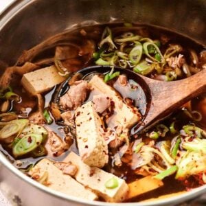 A pot of Korean kimchi stew with tofu, sliced beef, green onions, and spices is being stirred with a wooden spoon.