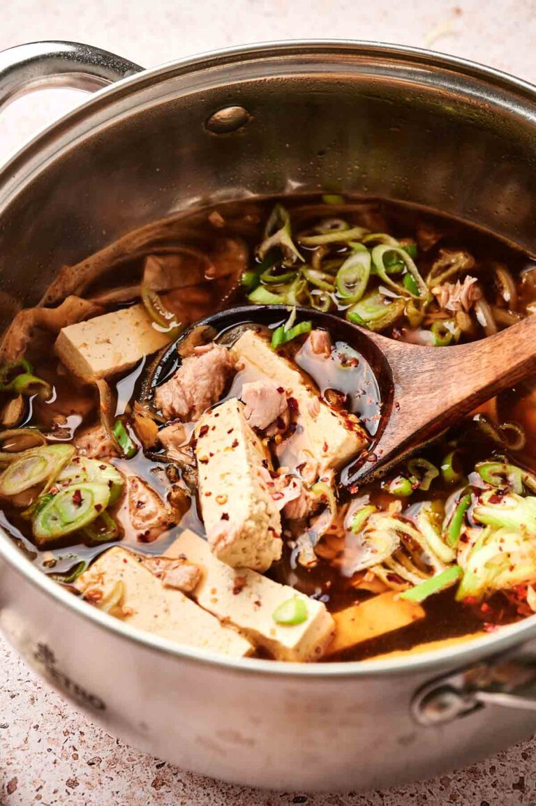 A pot of Korean kimchi stew with tofu, sliced beef, green onions, and spices is being stirred with a wooden spoon.