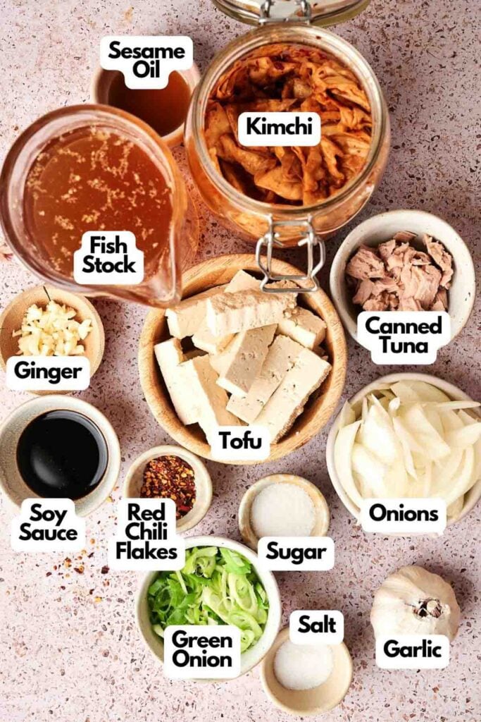 Ingredients for a delicious Kimchi Stew: kimchi, sesame oil, fish stock, canned tuna, tofu, onions, garlic, ginger, soy sauce, red chili flakes, sugar, salt, and green onion on a speckled surface.