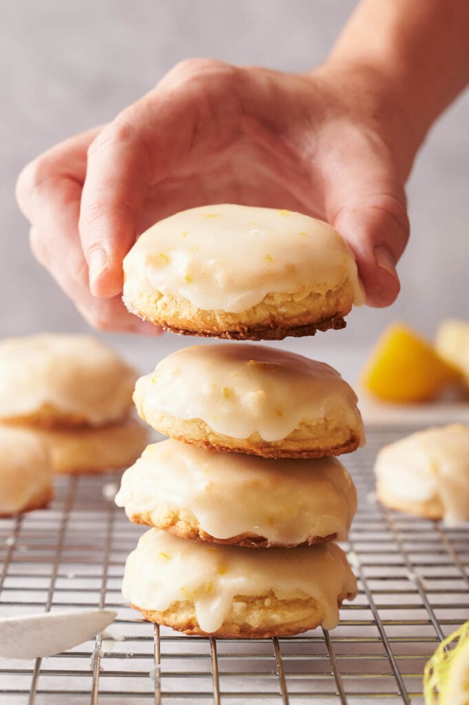 A hand holding a lemon-glazed cookie above a stack of three similar cookies on a wire rack, with lemon slices in the background.