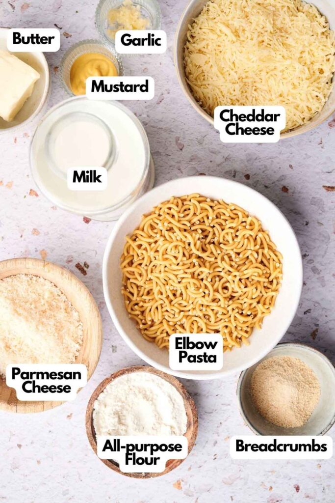 Ingredients for a pasta dish: bowls of elbow pasta, cheddar cheese, milk, breadcrumbs, all-purpose flour, parmesan cheese, butter, mustard, and garlic on a speckled surface.
