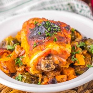 A roasted pork knuckle on a white plate, garnished with chopped parsley, sits atop a bed of roasted vegetables, including Brussels sprouts and sweet potatoes, reminiscent of the hearty goodness found in a Maple Dijon Chicken Sheet Pan dinner.